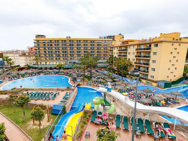 Duży hotel z wieloma basenami na zewnątrz, zjeżdżalniami wodnymi i leżakami pod pochmurnym niebem.