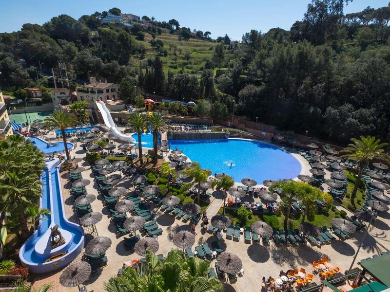 Duży basen hotelowy z zjeżdżalnią wodną, parasolami i zielonym otoczeniem.