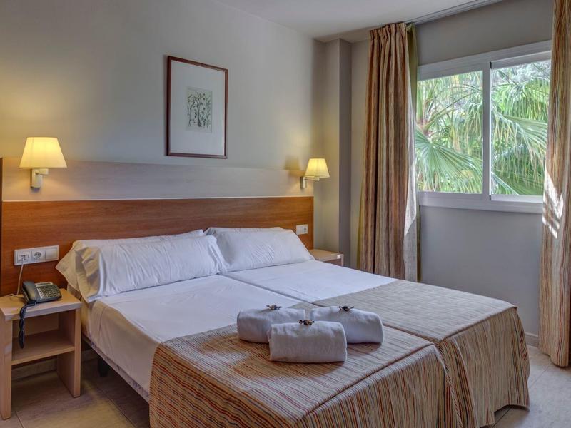 Jasny pokój hotelowy z podwójnym łóżkiem, stolikami nocnymi, lampami i oknem z widokiem na palmy.