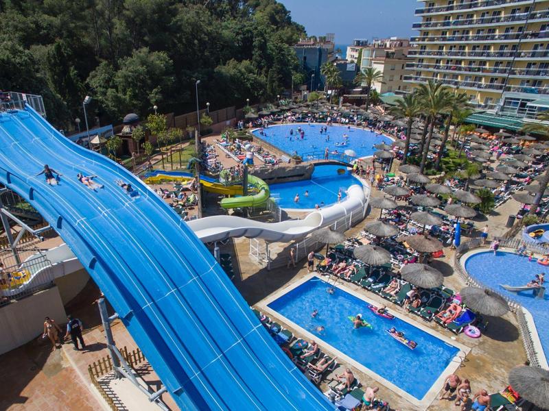 Duży hotel z wieloma basenami i zjeżdżalniami wodnymi otoczony leżakami i zielonymi drzewami.