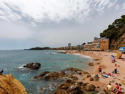 Plage avec rochers, personnes et bâtiments au bord de la mer sous un ciel nuageux.