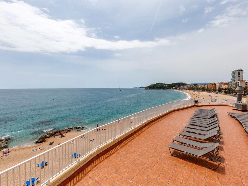 Terrasse ensoleillée avec transats offrant une vue sur une large plage de sable et la mer.