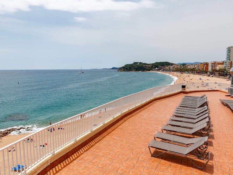 Plage avec mer calme, sable, chaises longues sur terrasse et immeubles au bord.