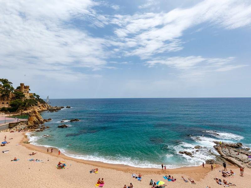 Plage de sable avec eau claire et personnes, entourée de rochers et végétation méditerranéenne.