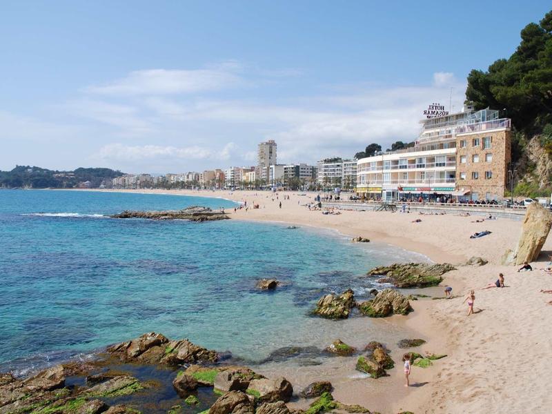 Plage avec eau claire, rochers et bâtiments au bord sous un ciel bleu.