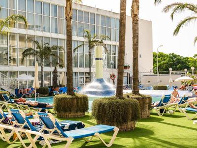 Hotel pool area with lounge chairs, umbrellas, and palm trees on green grass.