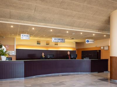 A modern hotel reception with a long front desk, signs, and two staff members.
