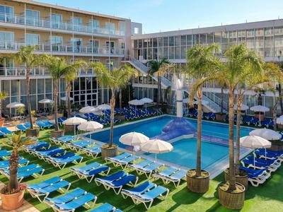 Hotel pool with sun loungers and palm trees in a modern hotel complex under clear sky.