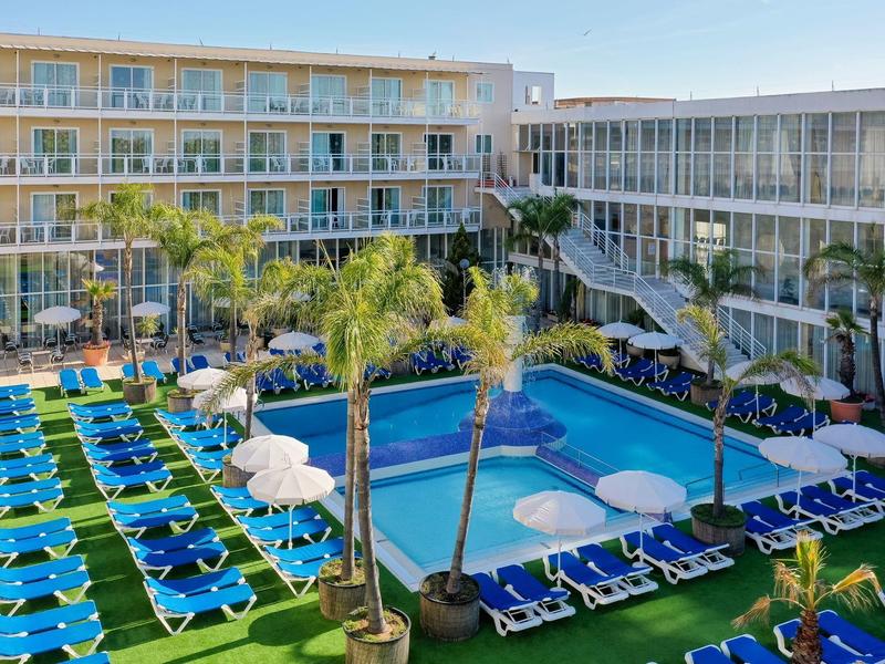 Hotel with outdoor pool surrounded by sun loungers and umbrellas under clear sky.