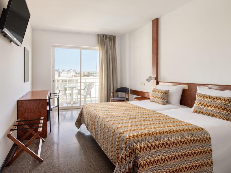 Bright hotel room with double bed, TV, desk, and balcony overlooking city.