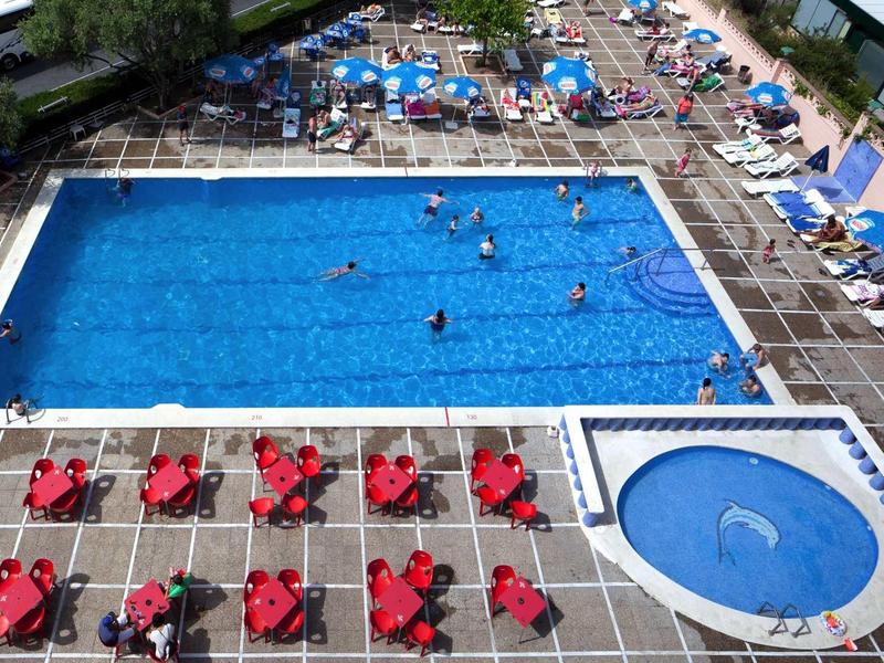 Outdoor pool with swimmers, surrounded by lounge chairs with red umbrellas.