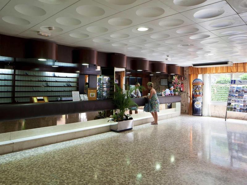 Hotel reception area with large counter, plants, and female guest.