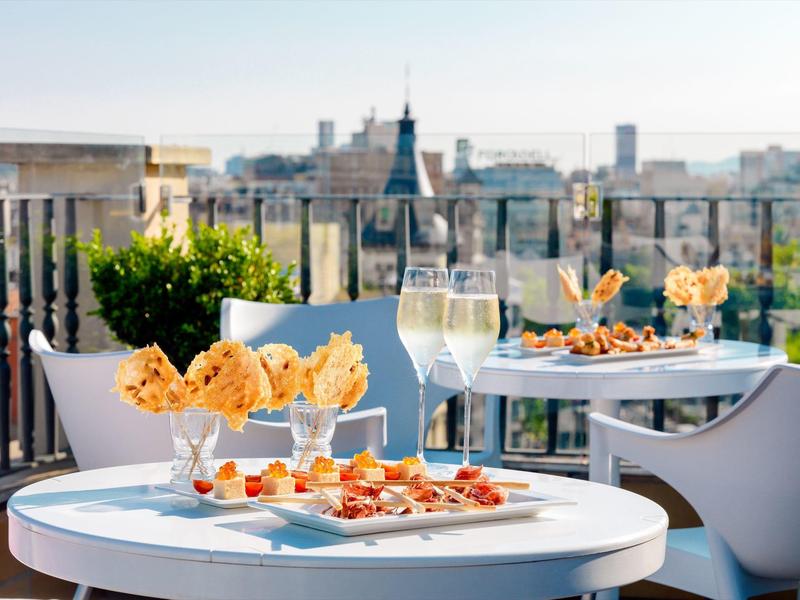 Tavoli bianchi con antipasti e due bicchieri di vino bianco su una terrazza con vista sulla città.