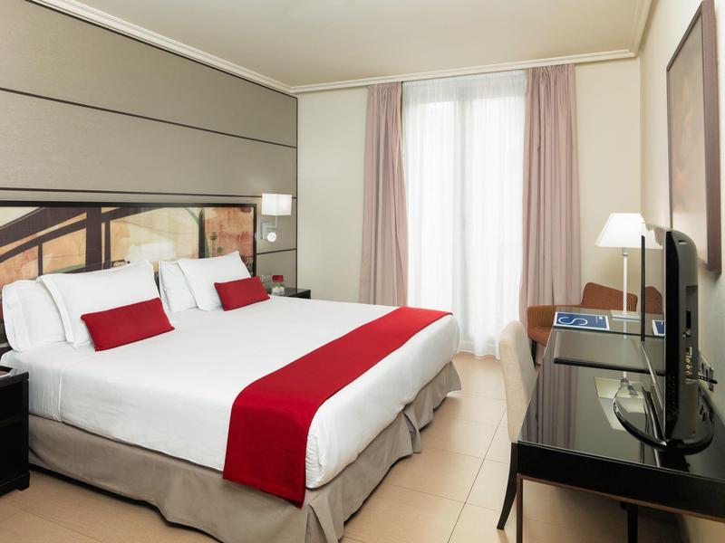 Camera d'hotel luminosa con letto grande, accenti rossi, scrivania e finestra con tende.