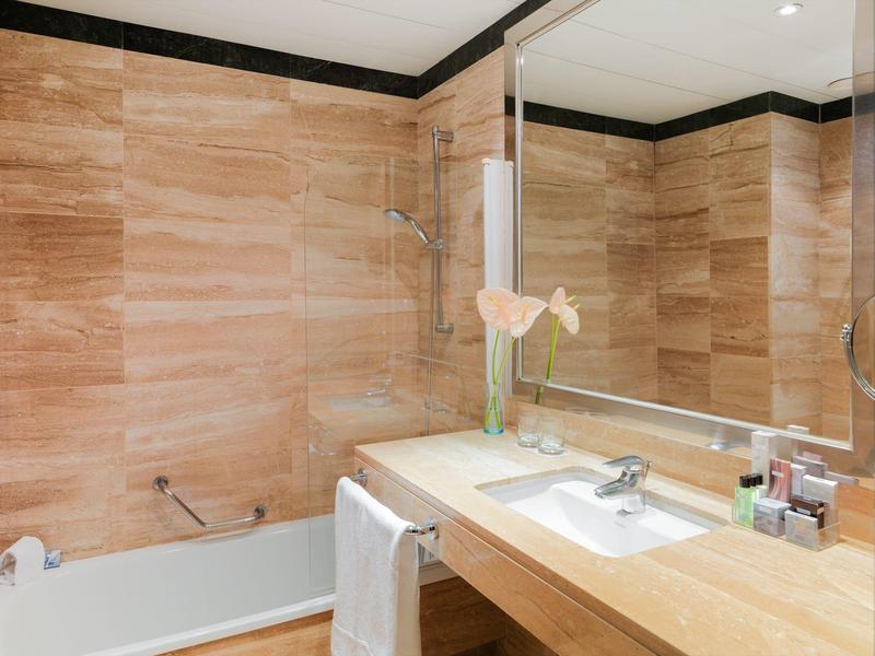 Bagno moderno con piastrelle beige, vasca, grande specchio e lavabo con articoli da bagno.