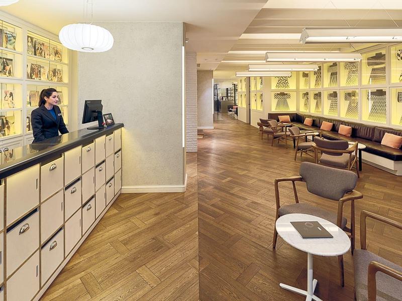 Hall d'hotel moderno con pavimento in legno, reception e area salotto con scaffali di libri.