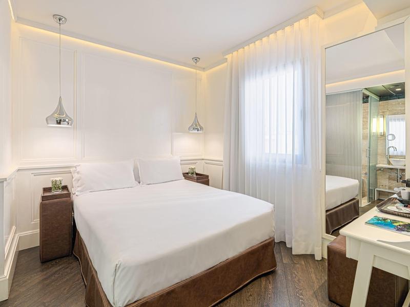 Camera d'hotel luminosa con letto matrimoniale, lenzuola bianche, tende e arredamento moderno.