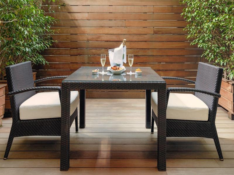 Set da pranzo da esterno con quattro sedie e un tavolo su un terrazzo in legno circondato da piante verdi.