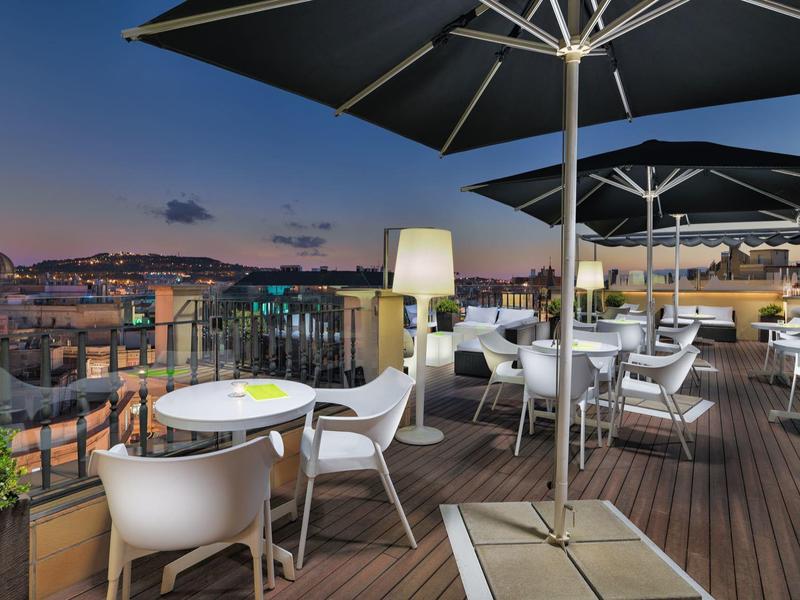 Terrazza panoramica con tavoli e sedie bianche, vista sulla città al crepuscolo sotto ombrelloni.