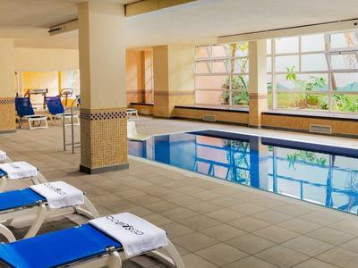 Piscina interior con tumbonas azules y grandes ventanas con vistas a áreas verdes.