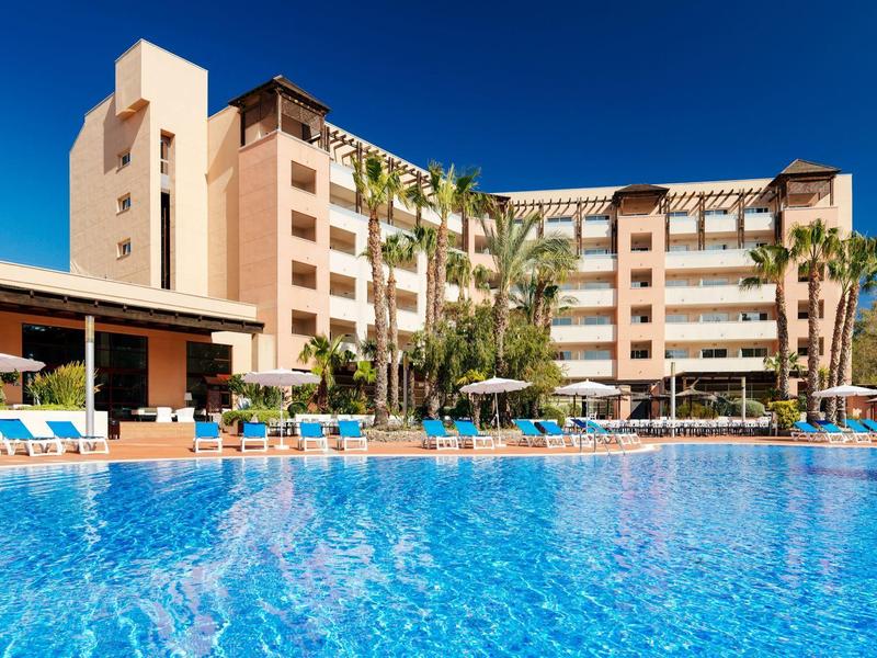 Edificio del hotel con palmeras y gran piscina bajo un cielo azul claro.