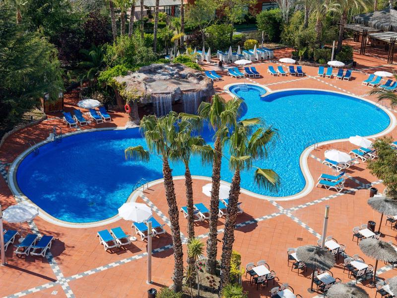 Piscina del hotel con palmeras, tumbonas azules y sombrillas en una terraza de piedra roja.