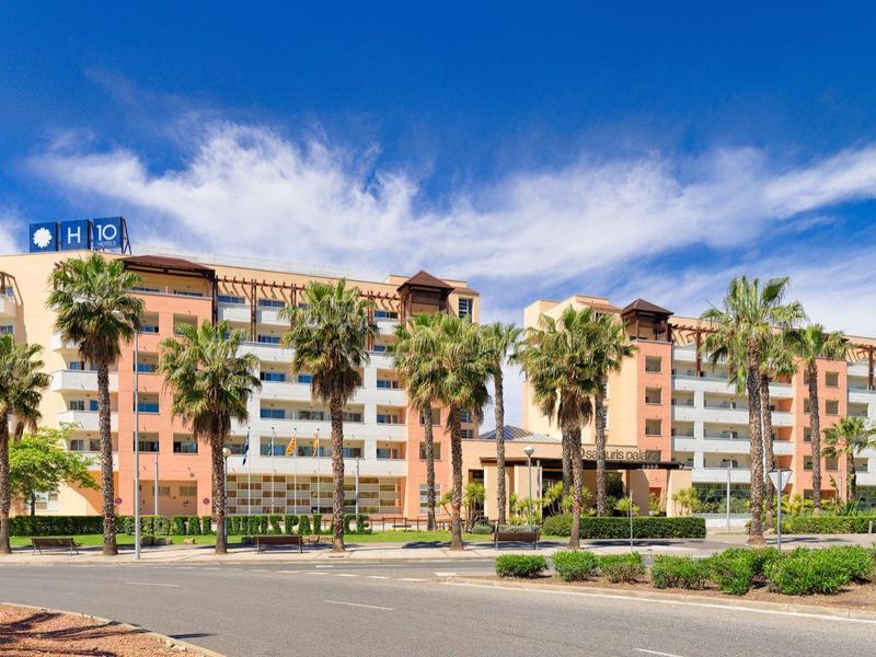 Gran edificio de hotel de varios pisos, rodeado de palmeras y césped verde bajo un cielo azul.