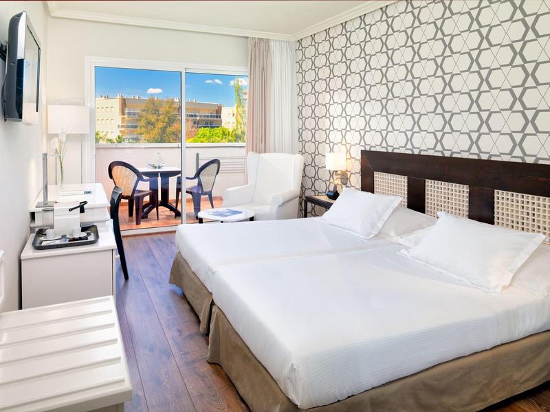 Habitación de hotel moderna con cama doble, sillas, balcón y vista a la ciudad de día.