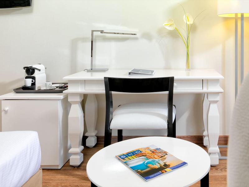 Área de trabajo moderna en hotel con escritorio blanco, silla, lámpara y cafetera en la habitación.