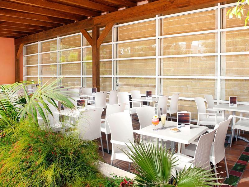 Área exterior cubierta de un hotel con sillas y mesas blancas, rodeada de plantas.