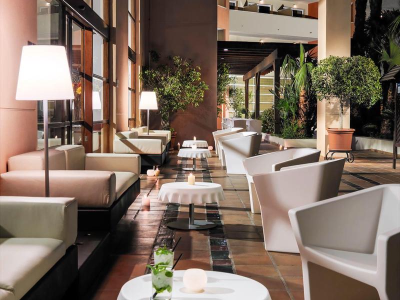 Acogedora zona de descanso en hotel con sillones blancos, mesas y plantas bajo vigas de madera.