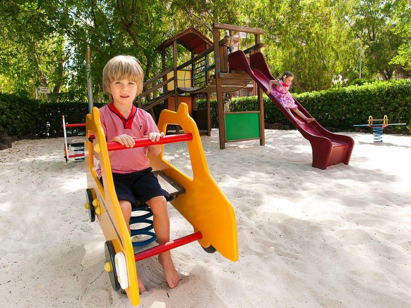 Niño jugando en un tobogán amarillo en un arenero soleado con estructura de escalada de fondo