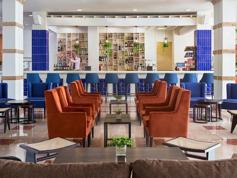 Lounge moderna de hotel con sillones naranjas, taburetes azules y pared decorativa.