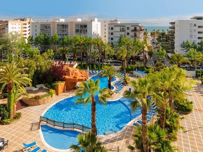 Resortzwembad met ligstoelen, palmbomen en witte gebouwen onder een blauwe lucht.