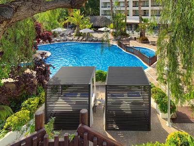 Zwembad van resort met ligstoelen, weelderig groen en een gebouw op meerdere verdiepingen op de achtergrond.