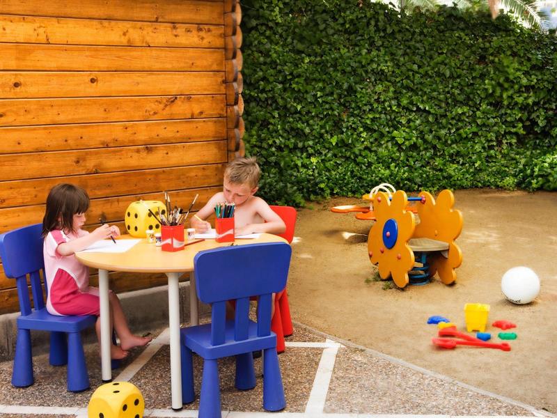 Kinderen spelen aan een klein buitentafeltje met speelgoed en kleurrijke stoelen op een zonnige dag.