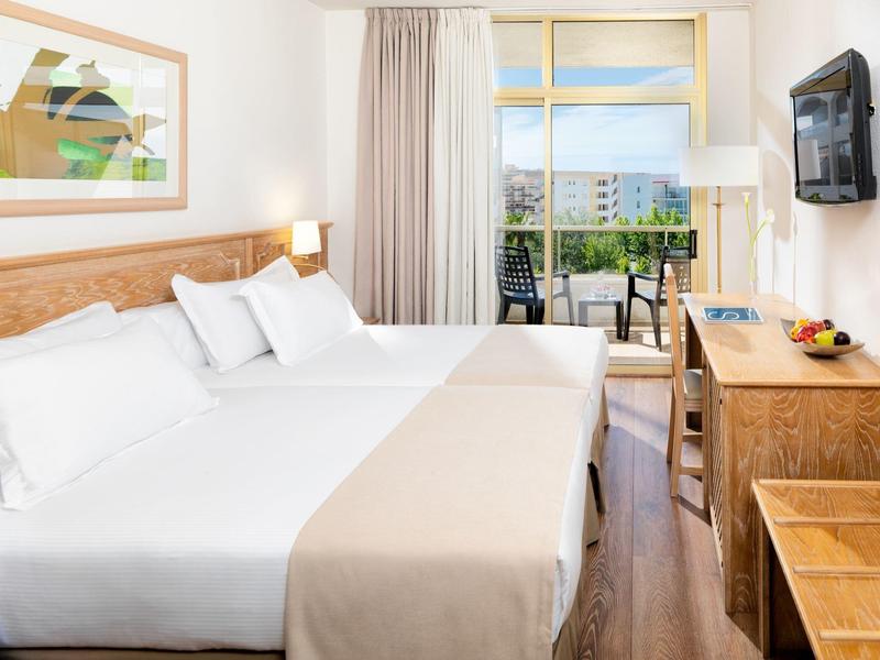 Lichte hotelkamer met twee bedden, houten meubels en balkon met uitzicht op stadsgebouwen.