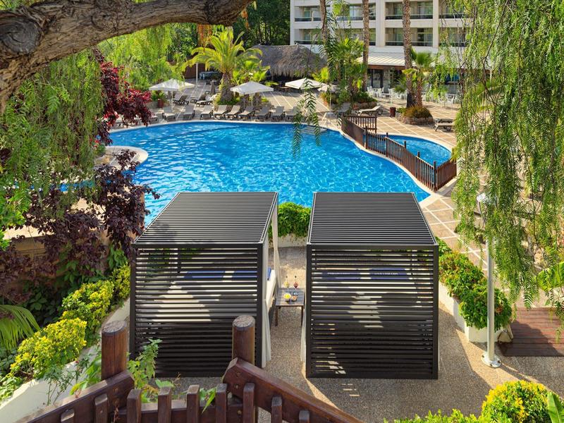Zwembad van resort met ligstoelen, weelderig groen en een gebouw op meerdere verdiepingen op de achtergrond.