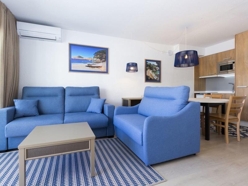 Salon lumineux avec canapés bleus, tapis et cuisine moderne en arrière-plan.