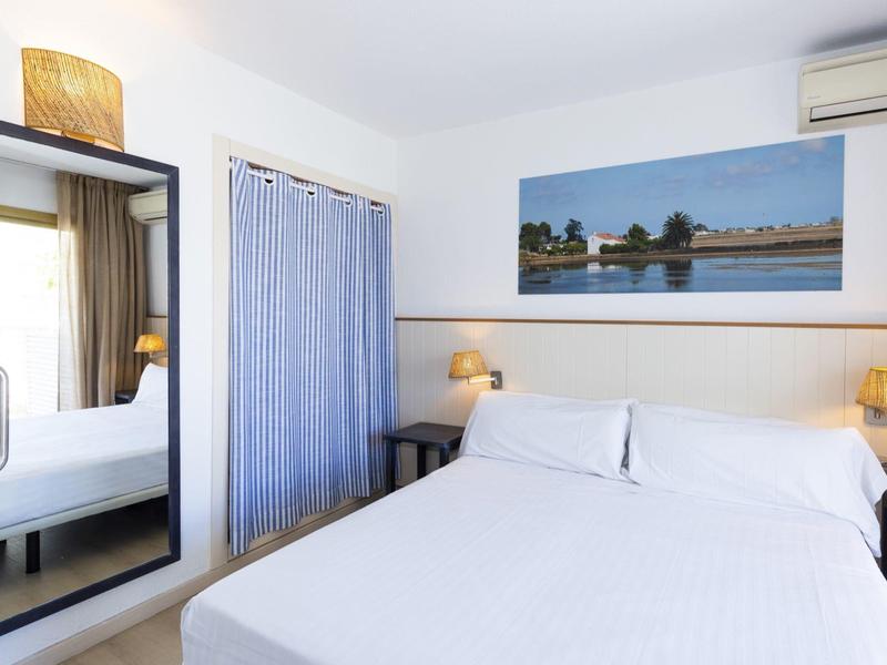 Chambre d'hôtel lumineuse avec grand miroir, rideau bleu et lit double avec linge blanc.