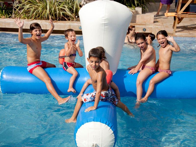 Enfants jouant dans une piscine sur un jouet flottant bleu et blanc, souriants et joyeux.