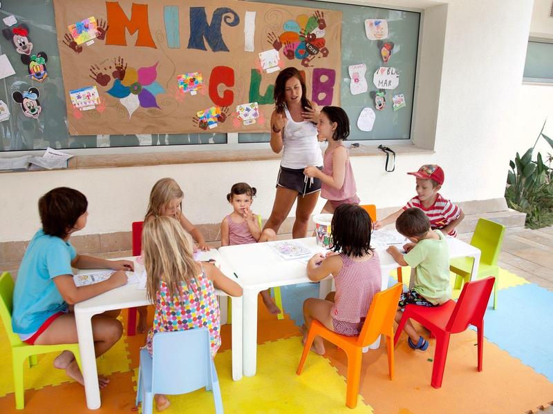 Enfants assis autour d'une table avec deux adultes dans une salle colorée.