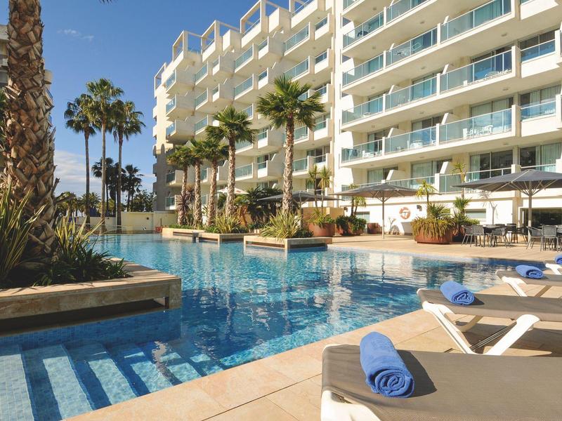 Piscine extérieure près d'un hôtel moderne avec chaises longues et palmiers.