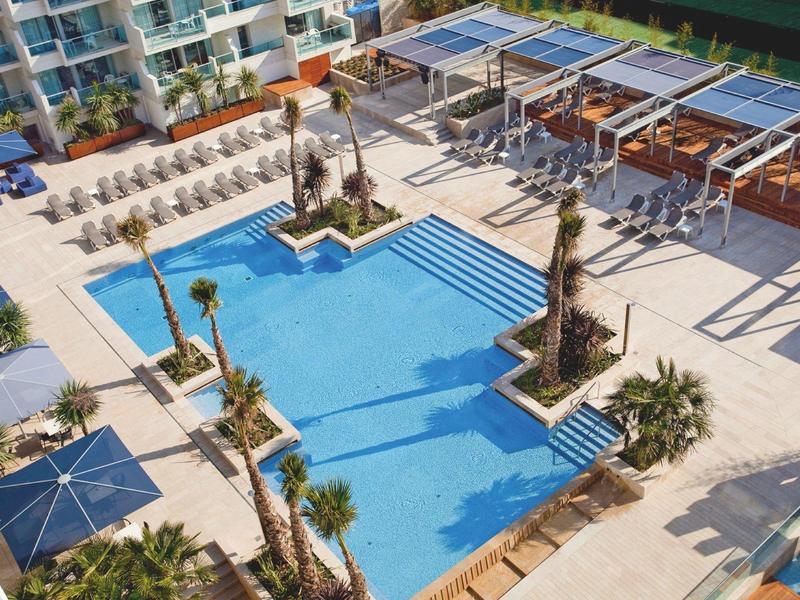 Piscine d'hôtel moderne avec statues, parasols bleus et chaises longues autour.