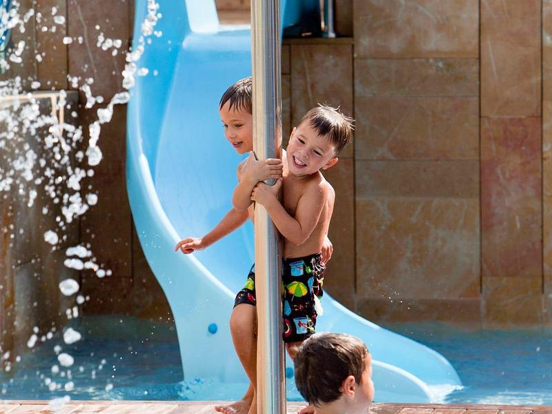 Deux garçons jouent près d'un toboggan bleu à côté d'une piscine avec jet d'eau.