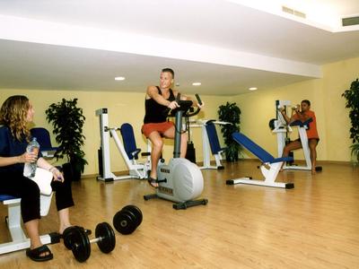 Drei Personen trainieren im hellen Fitnessraum mit verschiedenen Geräten und Hanteln.