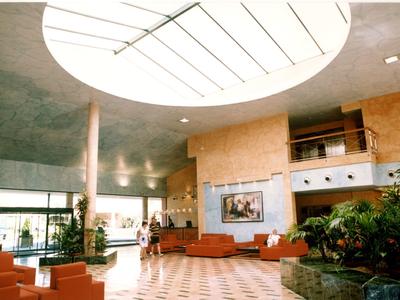 Spacious hotel lobby with circular skylight, orange seating, plants, and checkered floor.