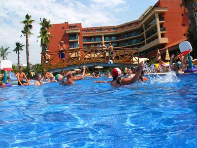 Menschen spielen Wasserball im Pool vor einem Hotel mit rotem Gebäude und einer Holzbrücke.