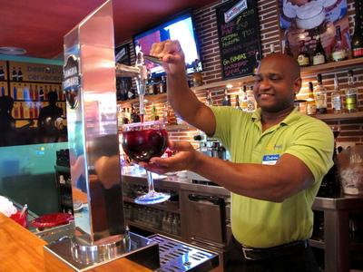 Bartender in green shirt holding a large glass of red wine behind the bar.