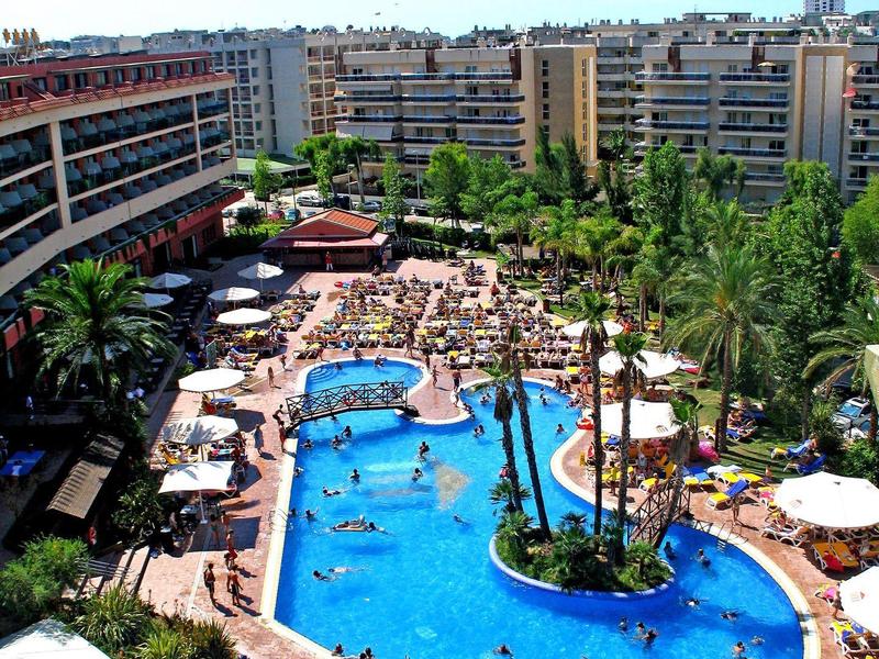 Resort with a large swimming pool surrounded by sun loungers and tall apartment buildings.